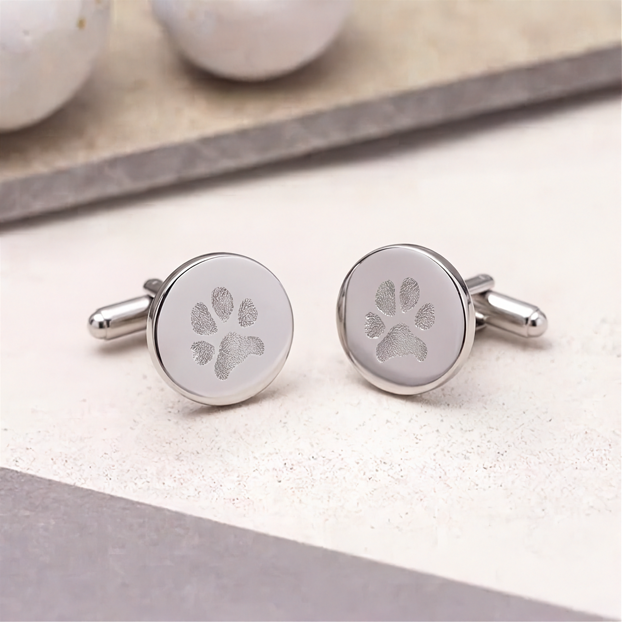 A close up of a pair of sterling silver, coin cufflinks with engraved paw prints, displayed on a light textured surface.  Custom men's jewelry from Atelier By DSGN.