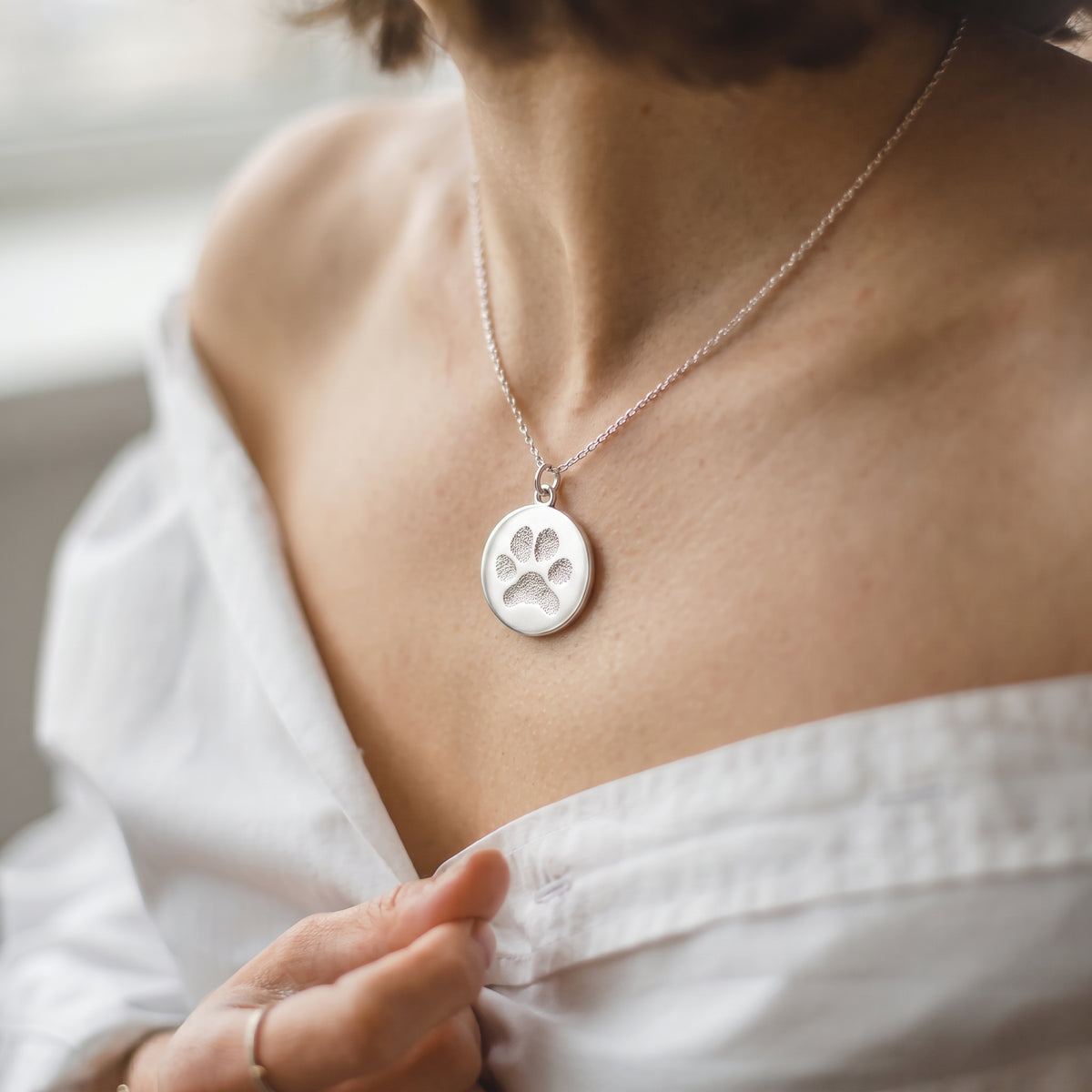 Female model in white low cut top holding custom engraved 925 sterling silver paw print coin pendant necklace at neckline, personalised pet paw print with  precision diamond-head engraved on polished round disc on delicate diamond-cut cable chain.  Atelier By Dsgn custom paw print pendant jewellery everyday wear gift for dog lovers, cat lovers, pet mum, pet memorial or rainbow bridge remembrance keepsake necklace.