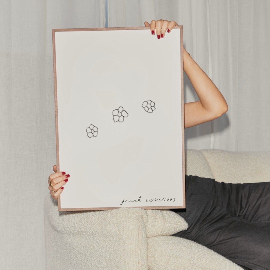 A person holds up a large minimalist framed art print featuring three small black flower-like, pet-nose-floral-line drawings on a white background, with a light wood frame. The person is seated on a cream textured sofa against a light curtain backdrop.  Meaningful art from Atelier By DSGN.