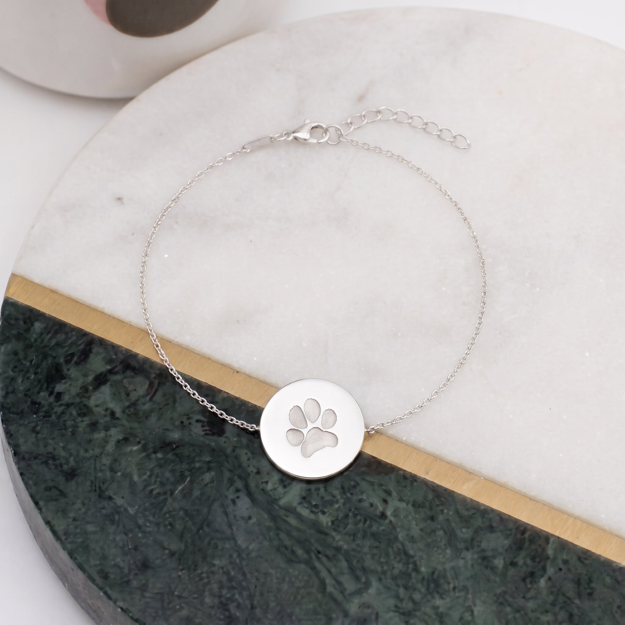 A delicate silver chain bracelet with a round disc pendant featuring a paw print engraving displayed on a white and green marble tray.  Custom jewelry from Atelier By DSGN