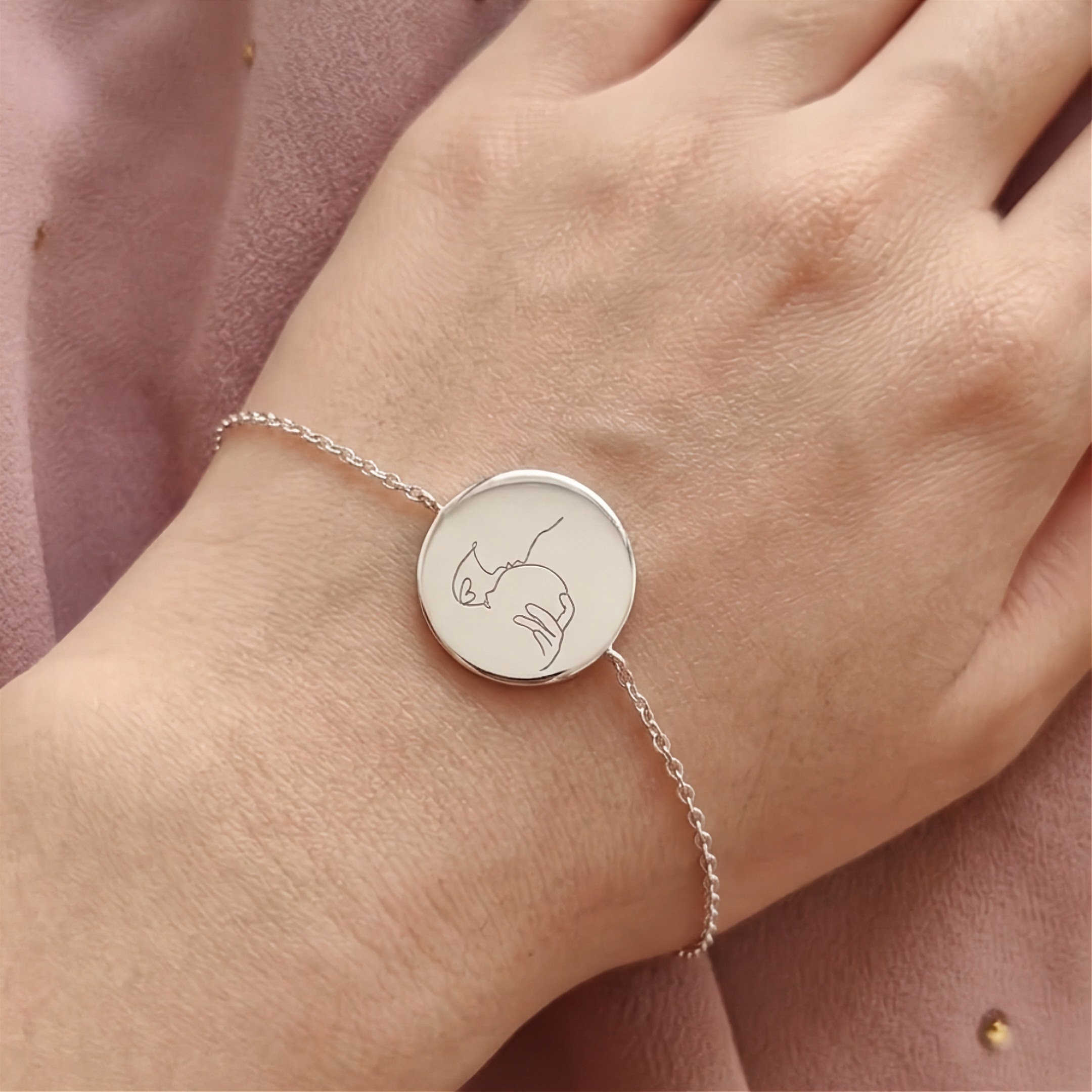 A delicate silver chain bracelet with a round silver disc pendant engraved with a minimalist line portrait drawing of a mom and baby shown worn on a person's wrist against a soft pink fabric background.  Meaningful custom jewelry from Atelier By DSGN
