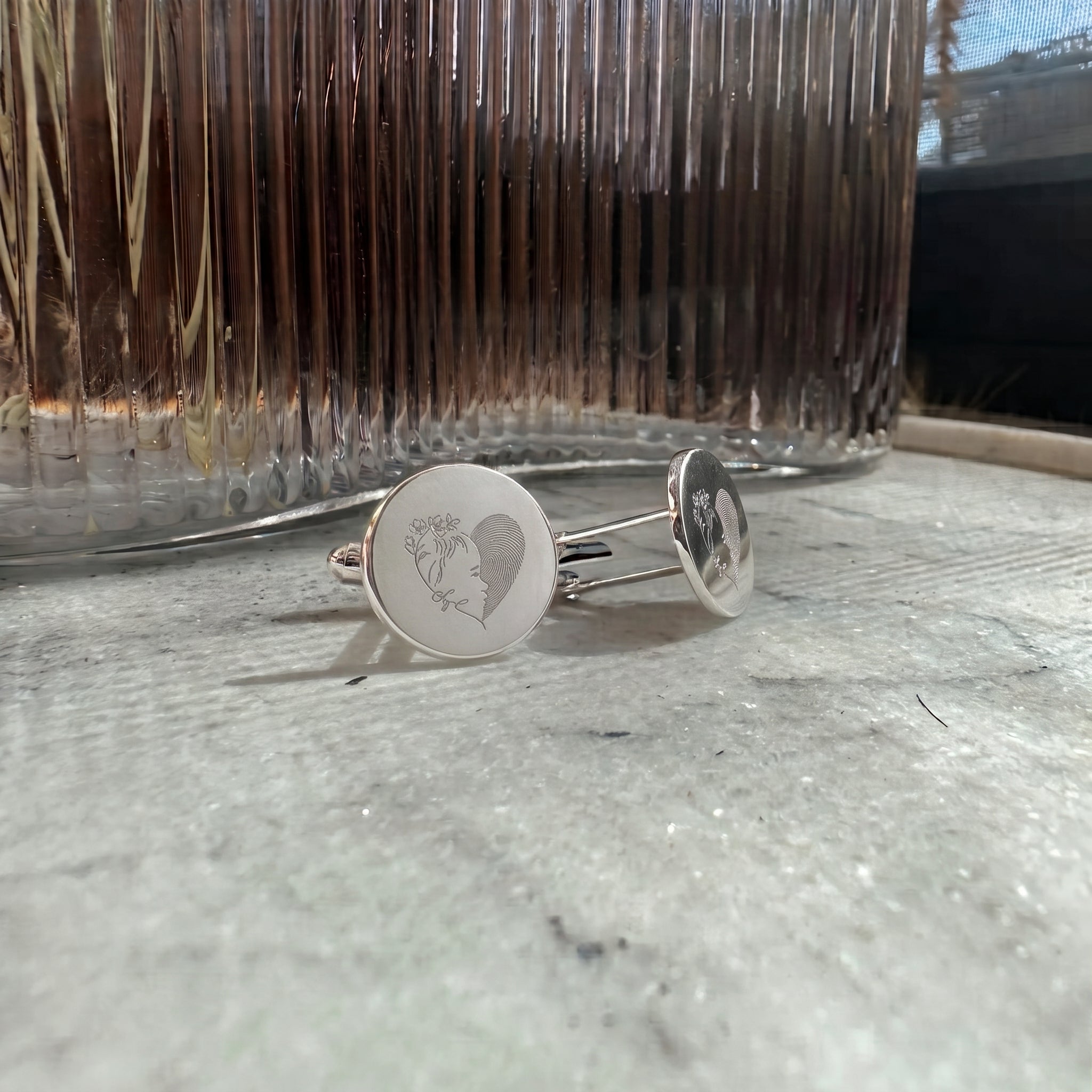 A close up of a pair of sterling silver cufflinks with engraved circular faces,  showing a single line portrait plus fingerprint engraved design in a heart motif.  Fingerprints are resting on a textured gray surface with a ribbed glass container in the background.  Custom jewelry from Atelier By DSGN.