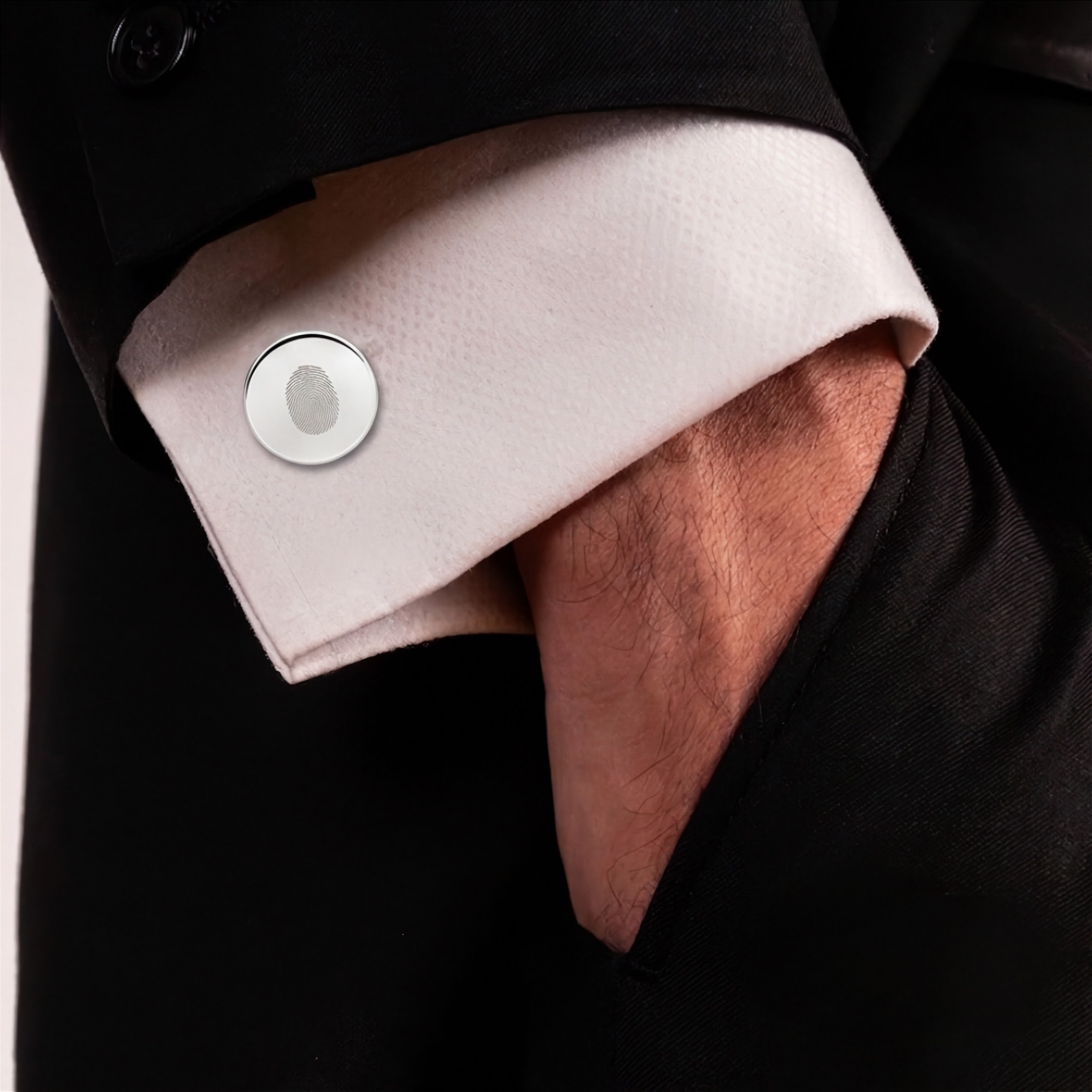 A close-up of a white dress shirt cuff with a single fingerprint engraved silver cufflink, worn under a black suit jacket. A hand is tucked into the jacket pocket.  Fingerprint jewelry from Atelier By DSGN