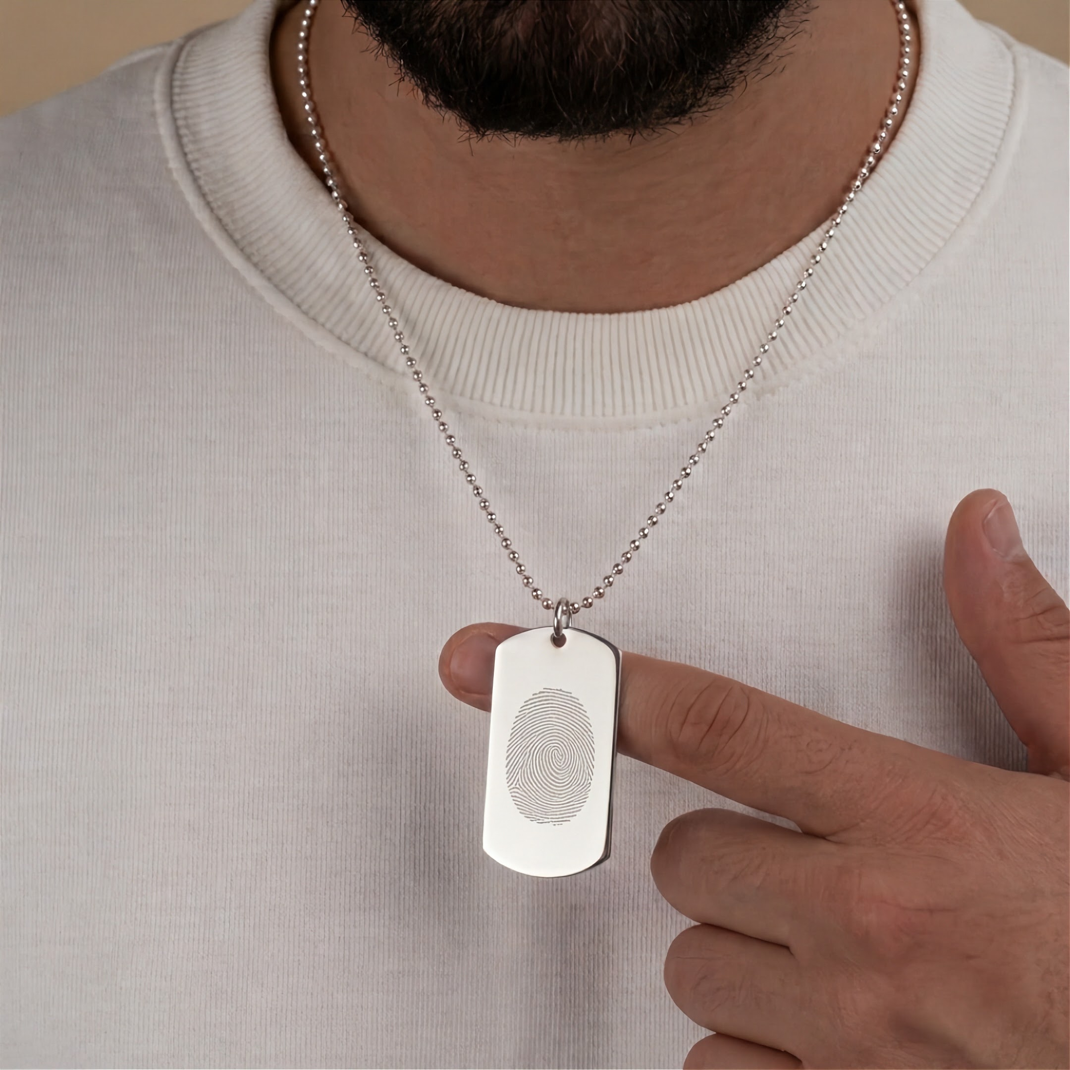 A silver fingerprint engraved dog tag necklace worn by a man, with his hand pointing to the fingerprint design on the pendant.  The pendant is on a beaded chain that is about chest length.  Custom jewelry from Atelier By DSGN.