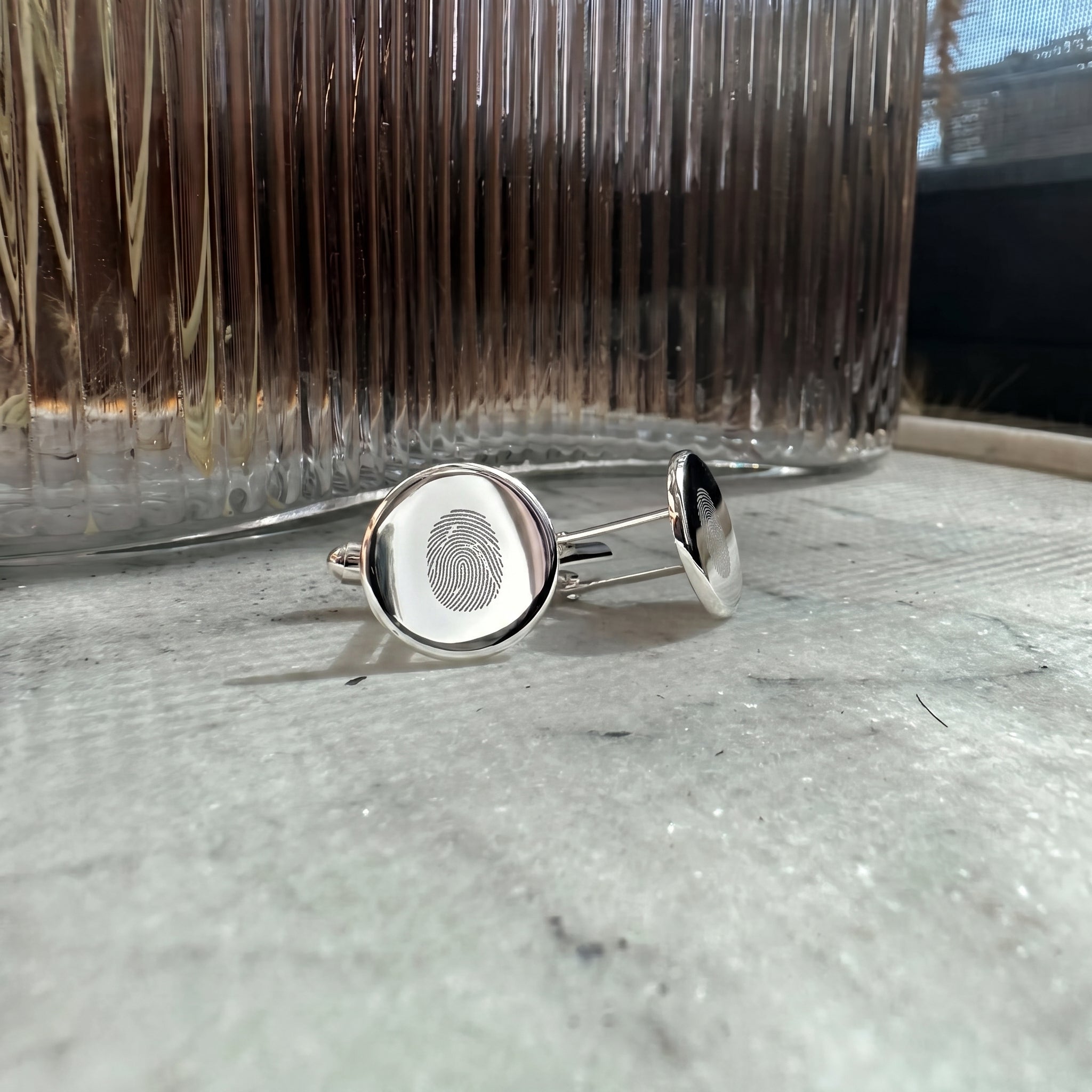 Single fingerprint engraved sterling silver cufflinks resting on a light grey marble table in front of a ribbed glass vase.  Custom men's jewelry from Atelier By DSGN.