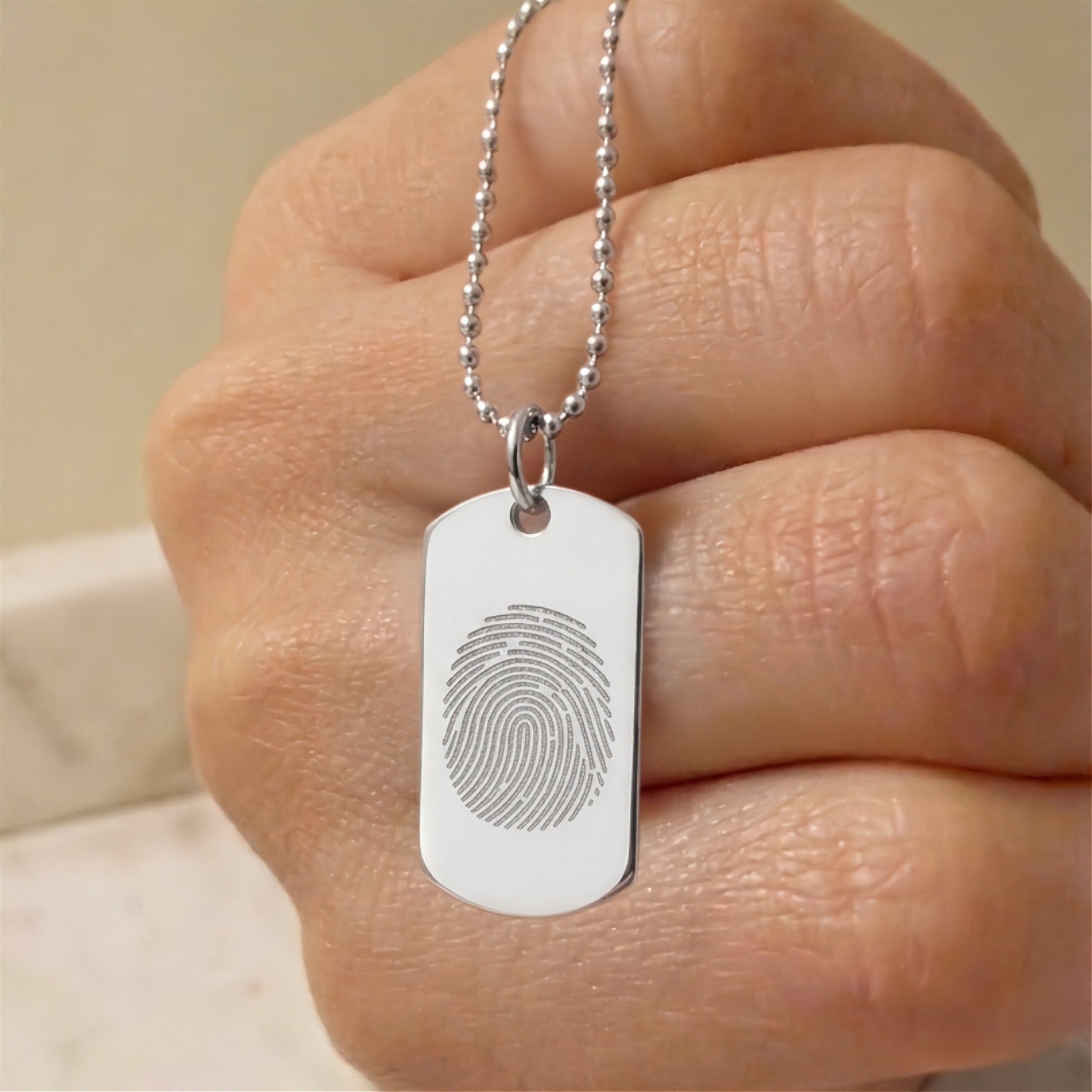 A close-up of a hand holding a silver ball-chain necklace with a dog tag pendant engraved with a single fingerprint design.  Custom meaningful jewelry from Atelier By DSGN.