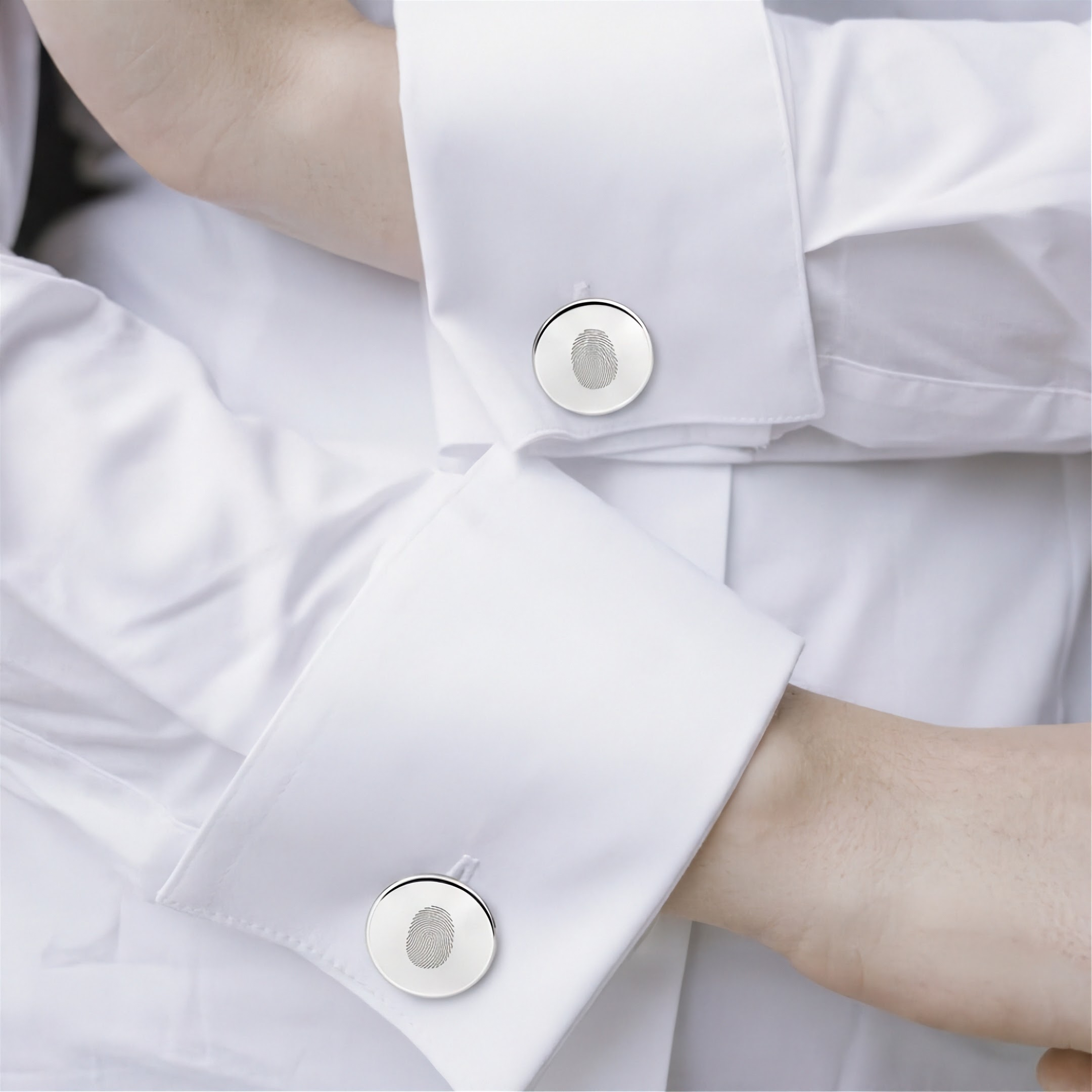 Close-up of a white dress shirt with two matching single sterling silver fingerprint engraved cufflinks and visible skin at the wrists.  Fingerprint jewelry from Atelier By DSGN