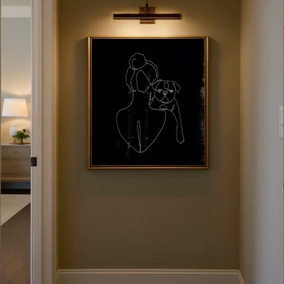 A framed black wall art print with white single line drawing of a lady and her bull dog, hanging on a warm beige hallway wall under a wall sconce. The image shows a portion of a home interior with a dark wood floor, white trim, and a hanging brass chandelier.  Customized line art from Atelier By DSGN.