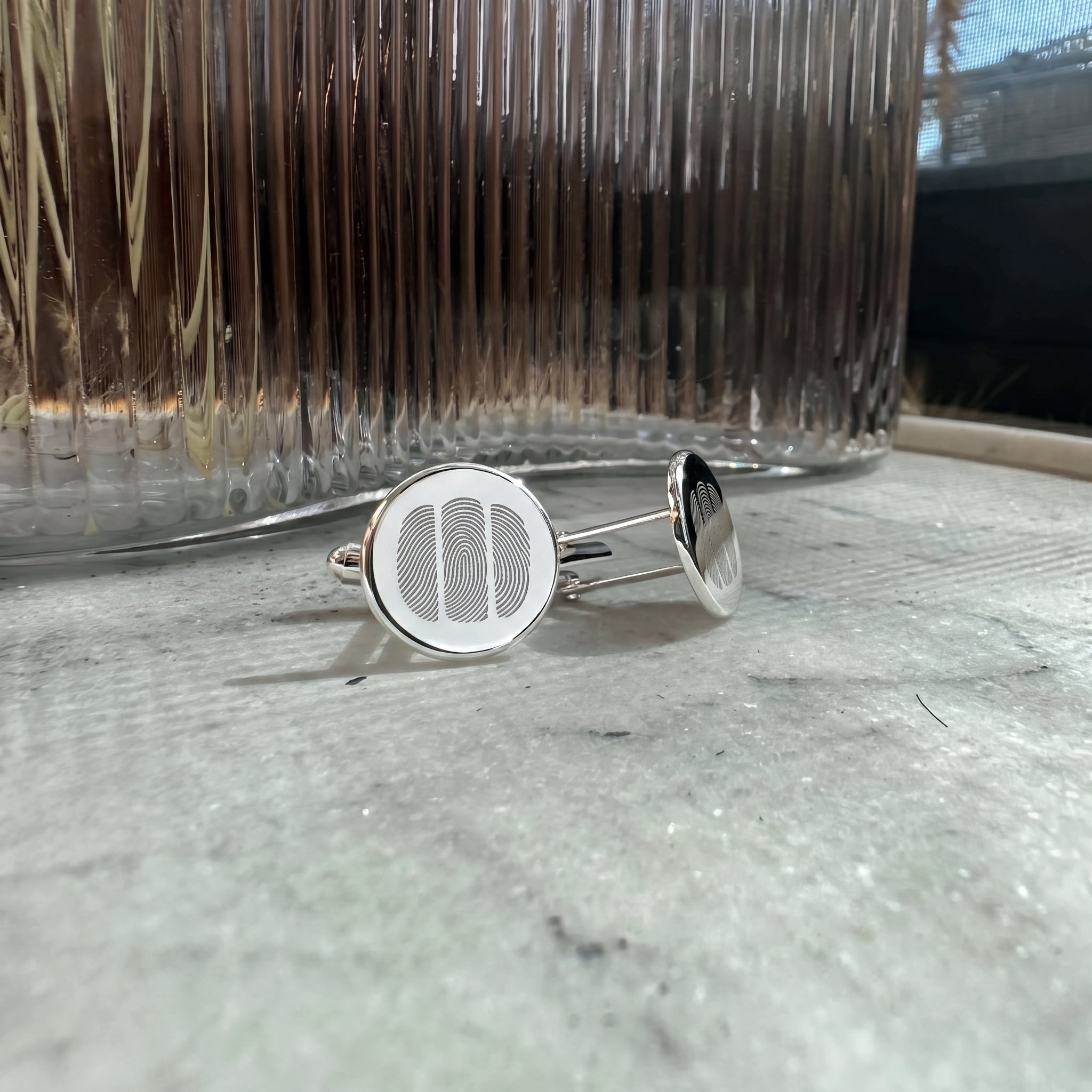 A pair of sterling siler coin circle cufflinks with a three fingerprint desgin engraved, resting on a light marble surface with a ribbed glass vase in the background.  Men's jewelry from Atelier By DSGN.