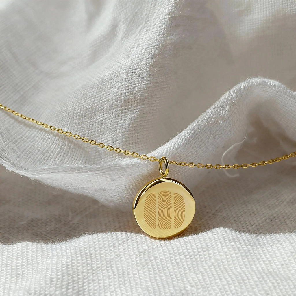 The Three Fingerprint Coin Necklace in 18K Gold Vermeil by Atelier By DSGN displayed on soft white fabric. The pendant showcases three diamond-engraved fingerprints on a polished gold surface, representing connection, love, and family in a luxurious, minimalist jewelry design.
