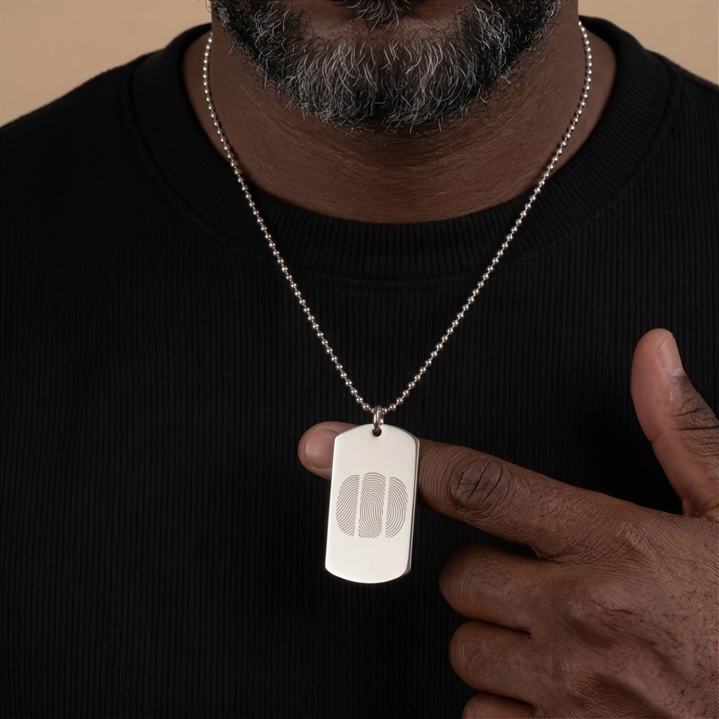 A close-up of a man wearing a silver ball-chain necklace with a rectangular dog tag pendant engraved with a three fingerprint design, paired with a black ribbed crewneck shirt against a warm beige background.  Custom jewelry from Atelier By DSGN.