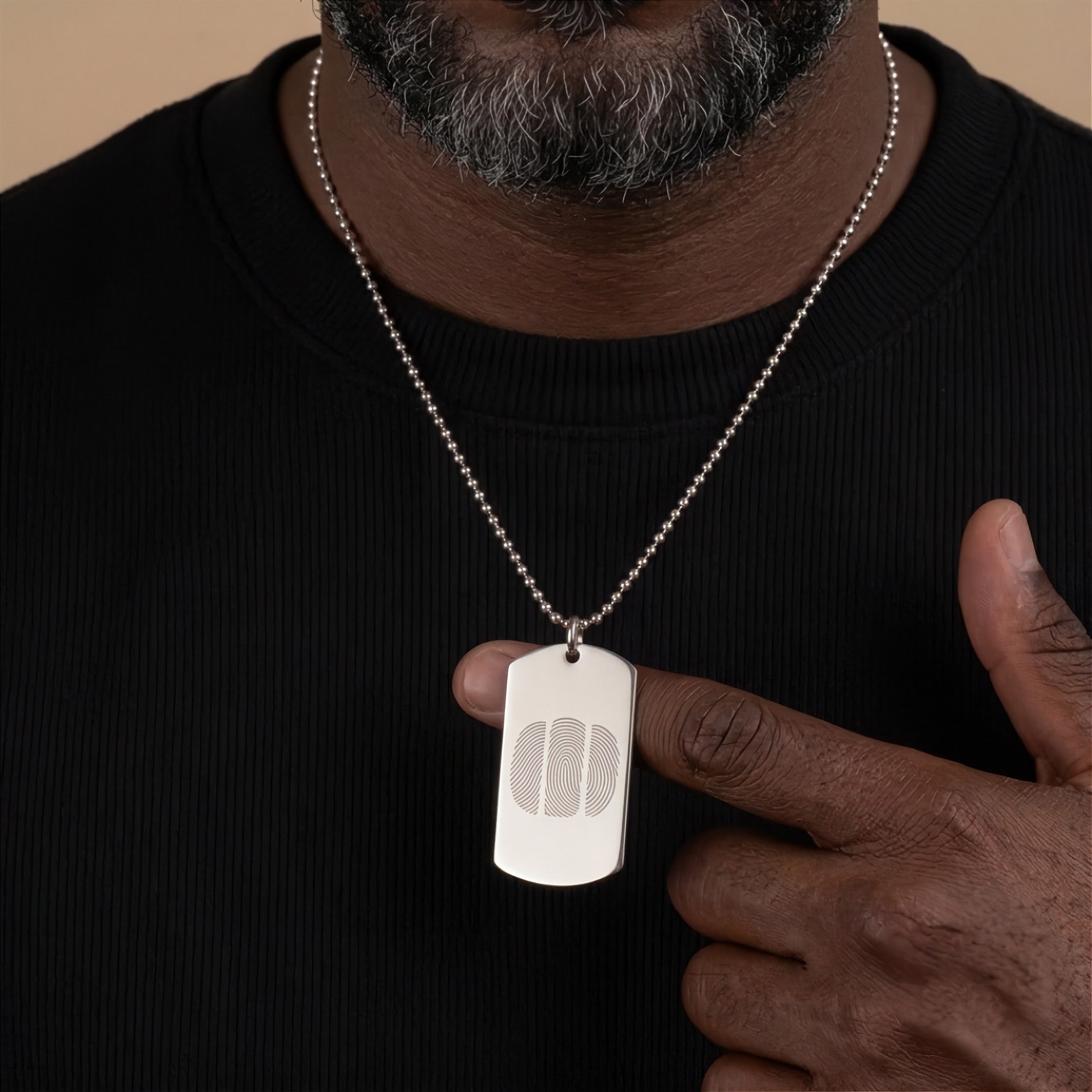 A close-up of a man wearing a silver ball-chain necklace with a rectangular dog tag pendant engraved with a three fingerprint design, paired with a black ribbed crewneck shirt against a warm beige background.  Custom jewelry from Atelier By DSGN.