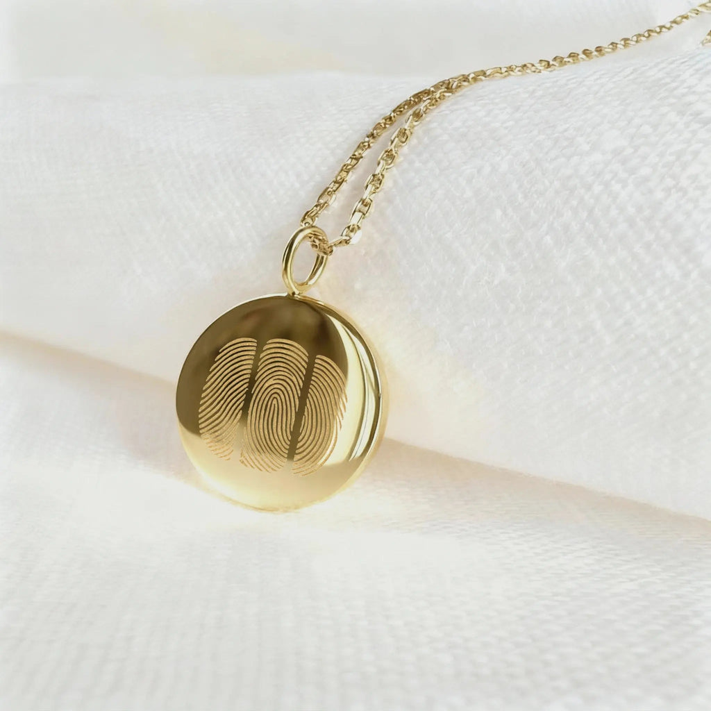 The Luxury Three Fingerprint Coin Necklace in 18K Gold Vermeil by Atelier By DSGN rests elegantly on a white fabric background. Featuring three detailed fingerprints engraved into a polished gold pendant, this handcrafted piece symbolizes love, memory, and connection in timeless luxury jewelry design.