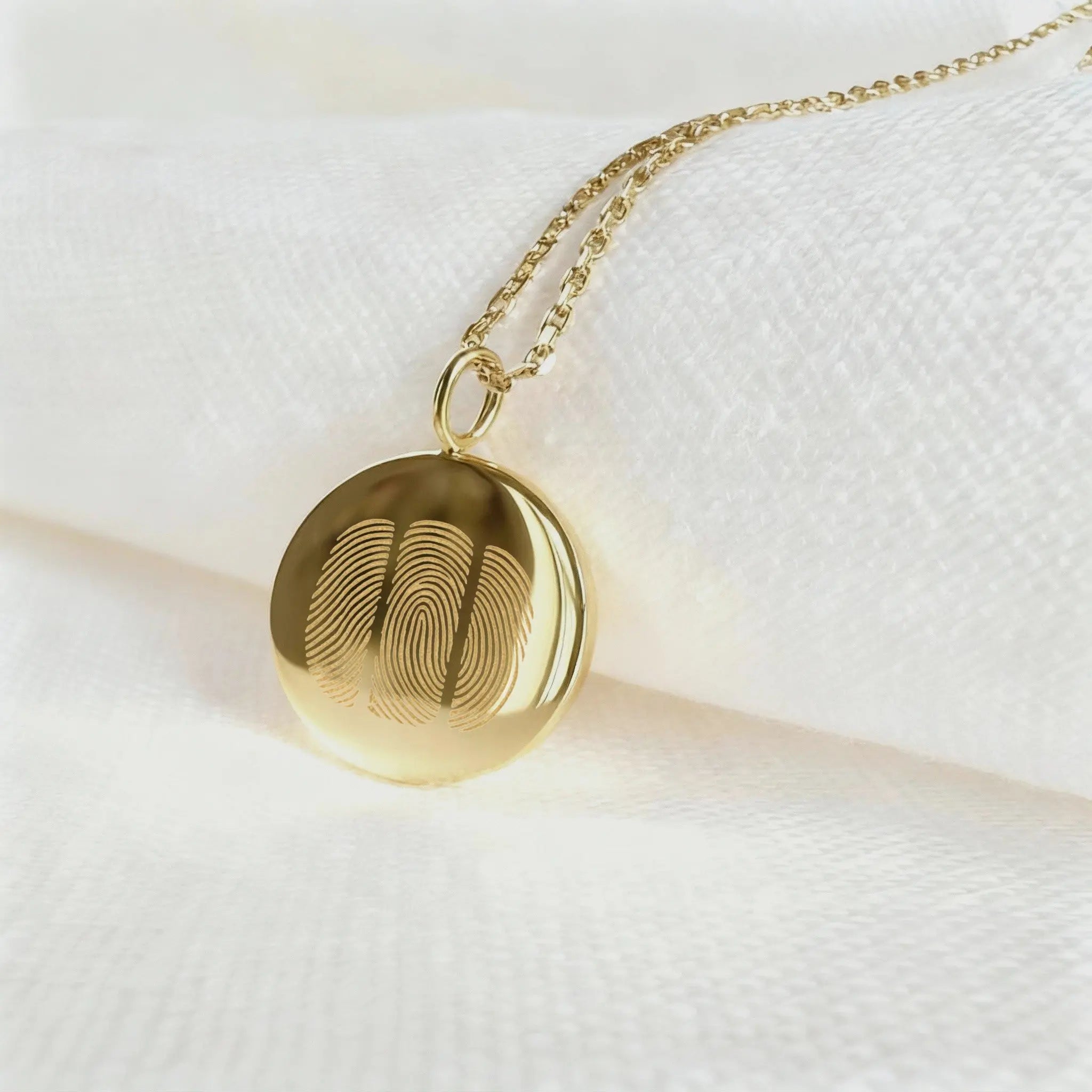 The Luxury Three Fingerprint Coin Necklace in 18K Gold Vermeil by Atelier By DSGN rests elegantly on a white fabric background. Featuring three detailed fingerprints engraved into a polished gold pendant, this handcrafted piece symbolizes love, memory, and connection in timeless luxury jewelry design.