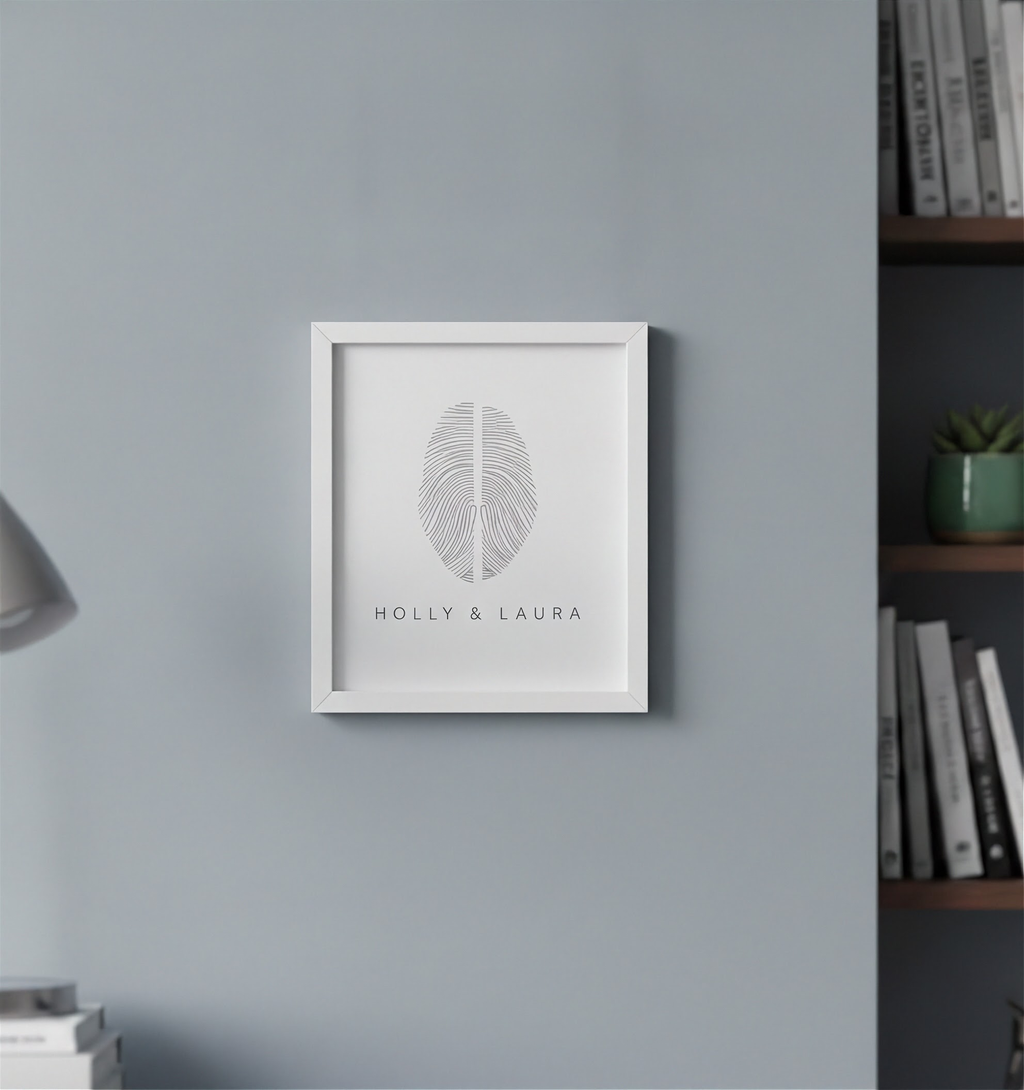 A framed personalized fingerprint artwork featuring a two fingerprint design and the names 'HOLLY & LAURA' in a white frame, mounted on a light gray wall in the study.  Custom two fingerprint art by Atelier By DSGN.