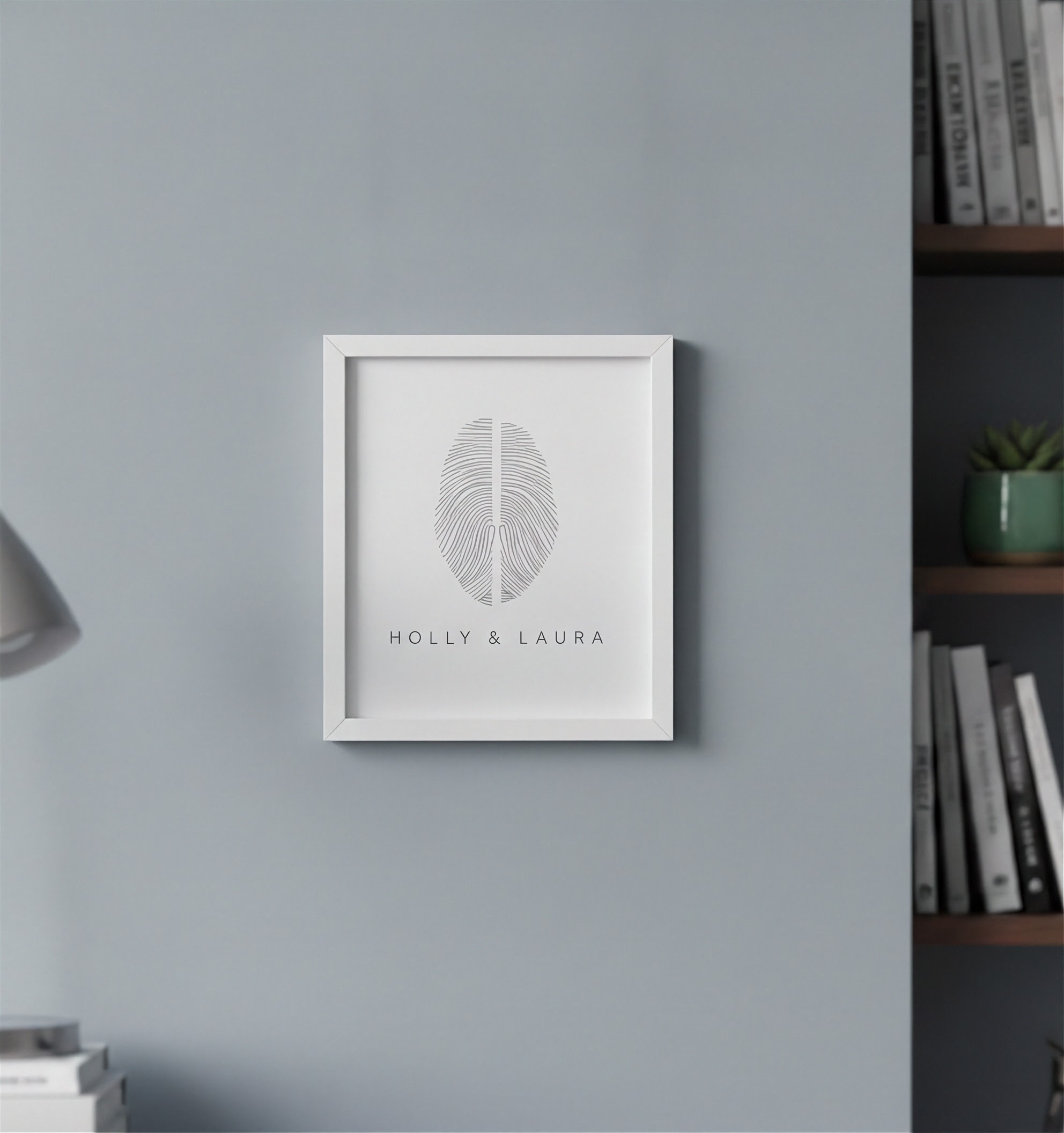 A framed personalized fingerprint artwork featuring a two fingerprint design and the names 'HOLLY & LAURA' in a white frame, mounted on a light gray wall in the study.  Custom two fingerprint art by Atelier By DSGN.
