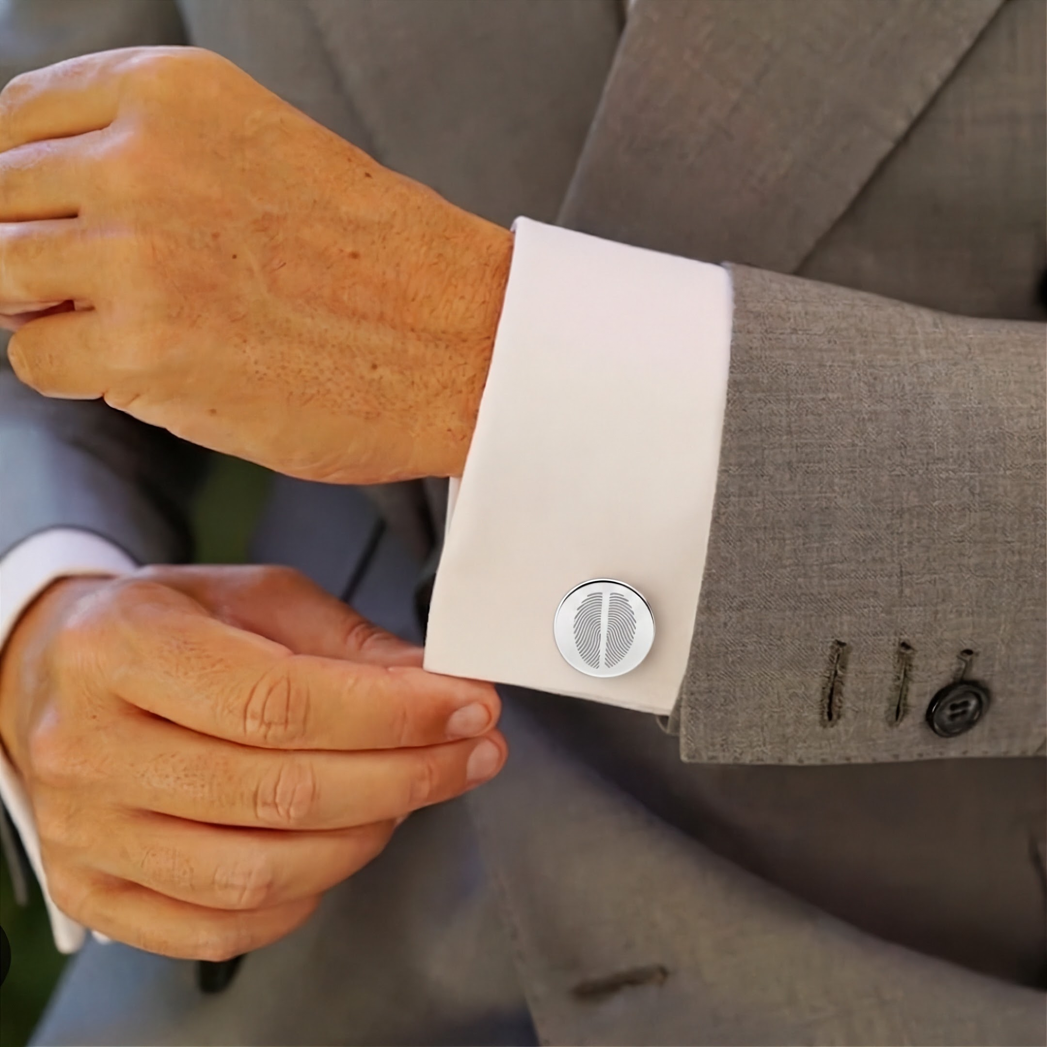 A close-up of a man's hands adjusting a white dress shirt cuff with a two fingerprint engraved round silver coin cufflink, worn with a gray suit jacket.  Meaningful men's jewelry from Atelier By DSGN.
